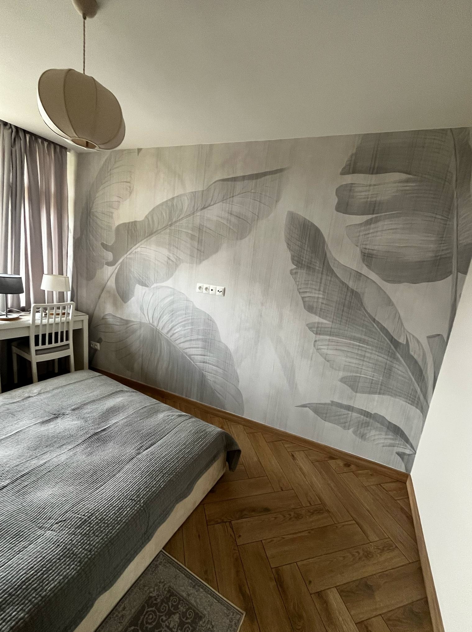 Sypialnia z fototapetą w liście, parkietem w jodełkę i minimalistycznym biurkiem. Lampa sufitowa w kształcie kuli. Stonowana kolorystyka, nowoczesny design.