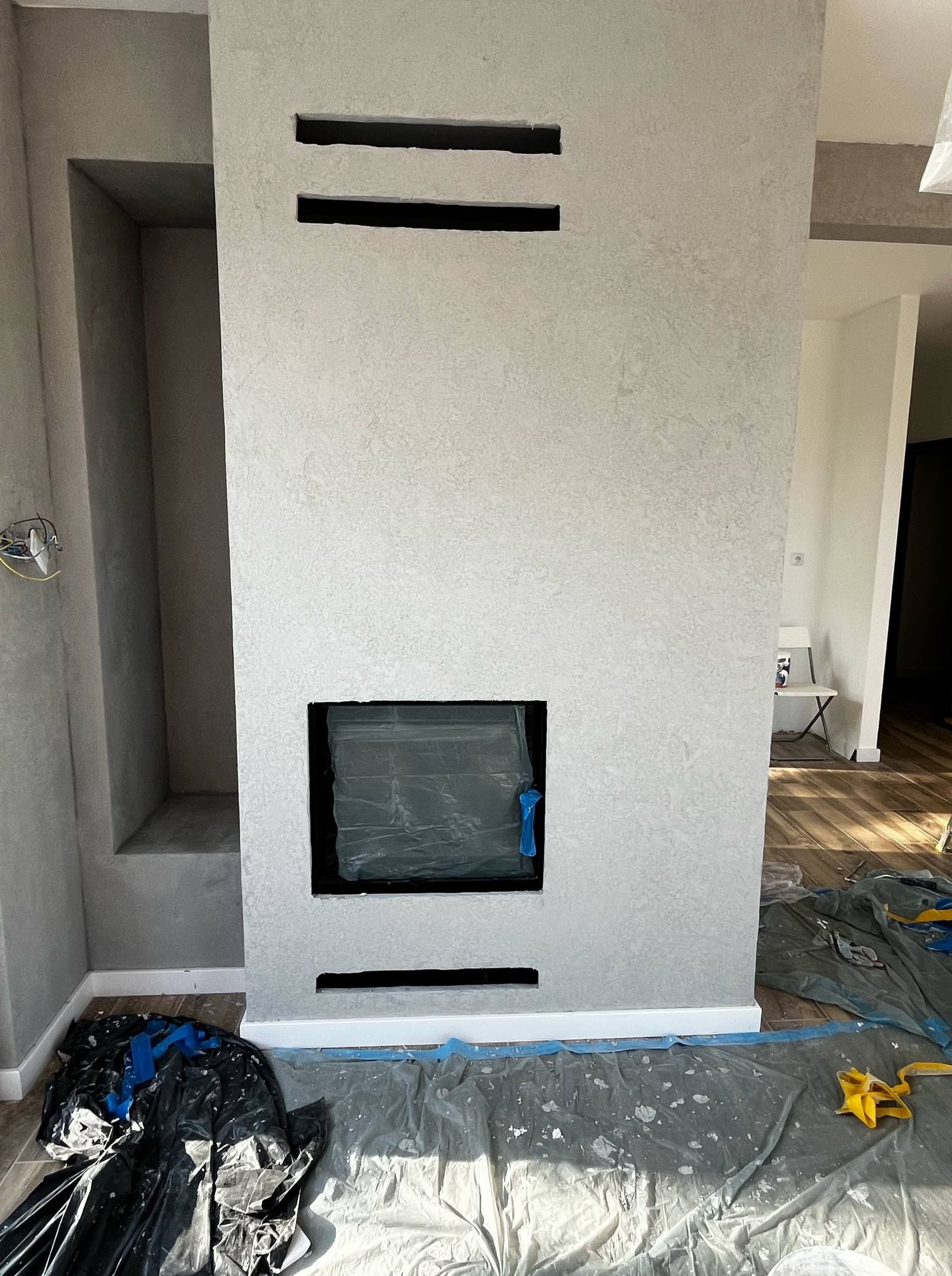 Ściana z szarą, dekoracyjną strukturą, z wnęką i otworem na kominek zabezpieczonym folią. Podłoga zabezpieczona folią malarską.