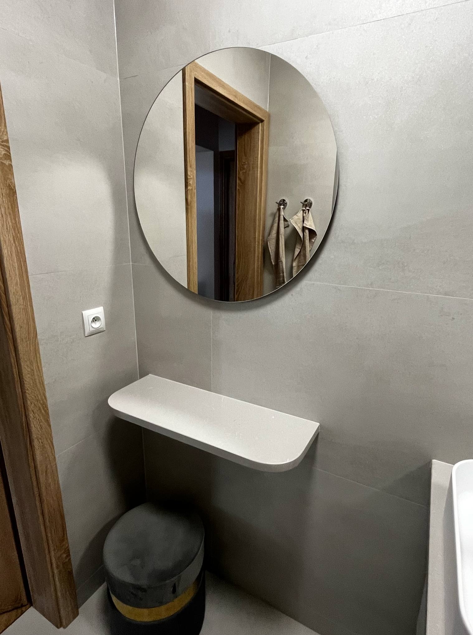 Szara łazienka z okrągłym lustrem, półką i stołkiem. Widoczne płytki na ścianach, drewniana futryna drzwi i fragment umywalki. Minimalistyczny design, stonowana kolorystyka.