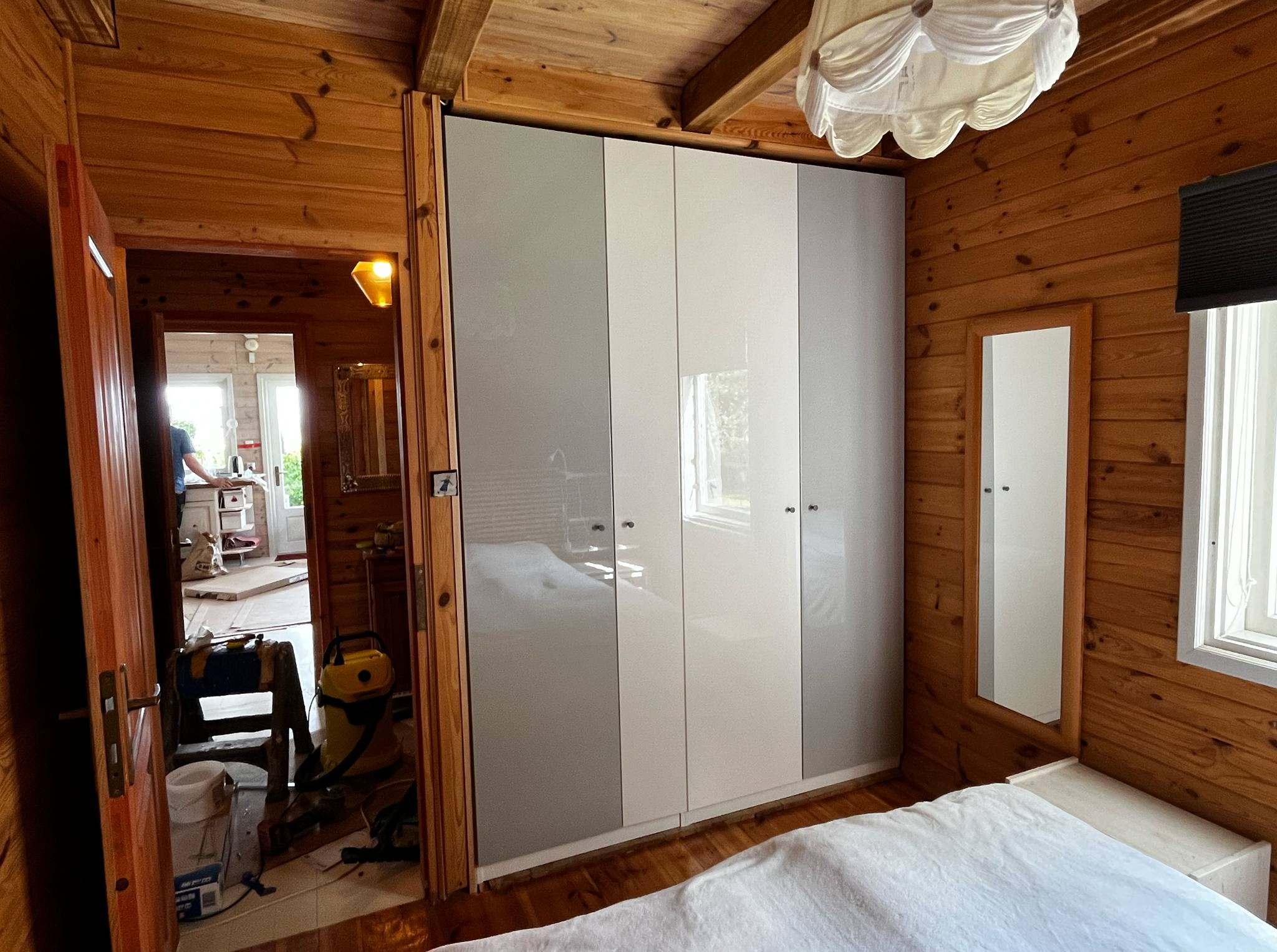 Jasny pokój z drewnianą boazerią i szafą w odcieniach szarości. W tle widoczne narzędzia i materiały budowlane, sugerujące trwające prace wykończeniowe.