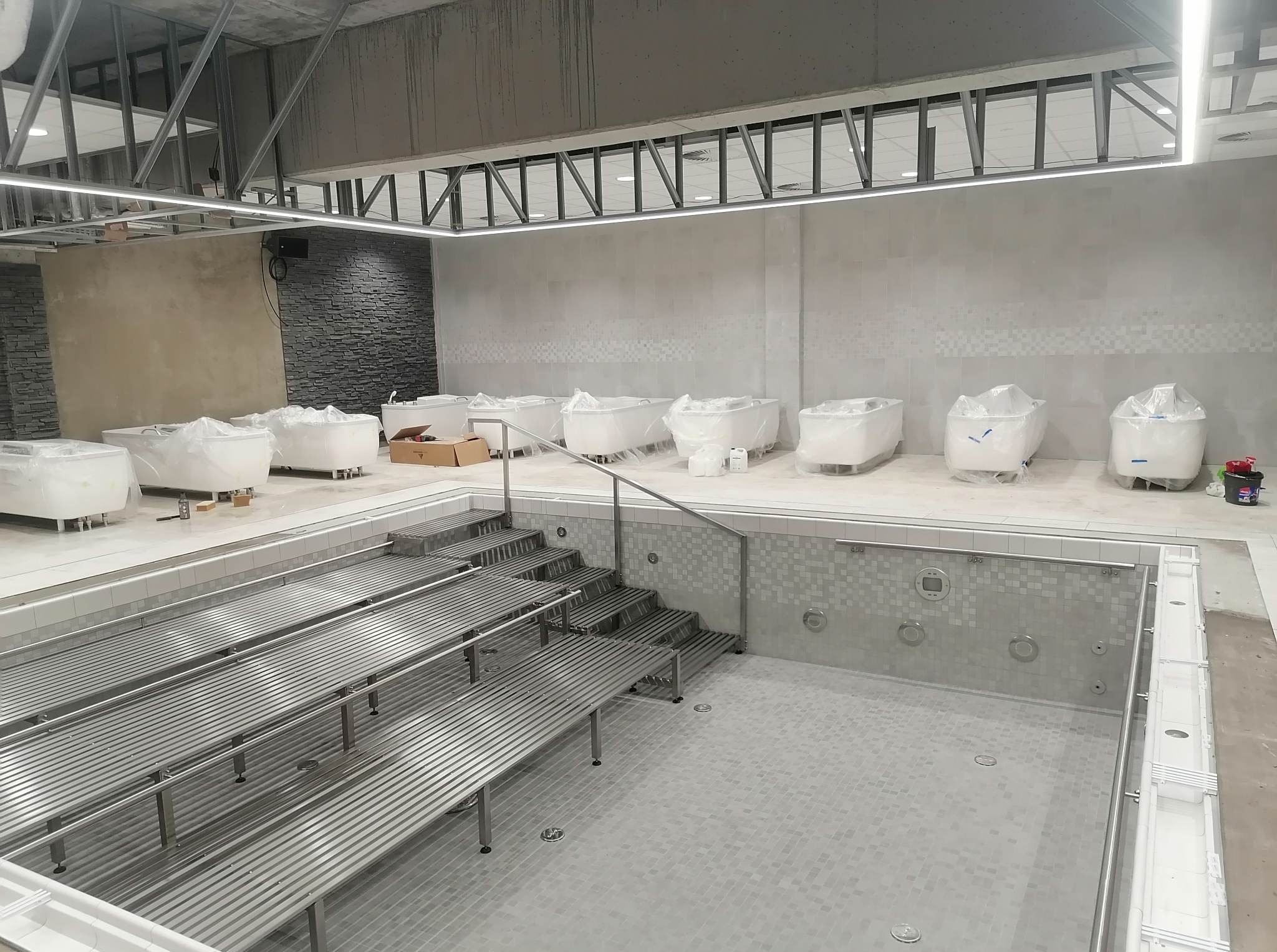 Wnętrze budowanego basenu z metalowymi leżankami i schodami, przy ścianach wanny jacuzzi zabezpieczone folią. Widoczna surowa konstrukcja sufitu.