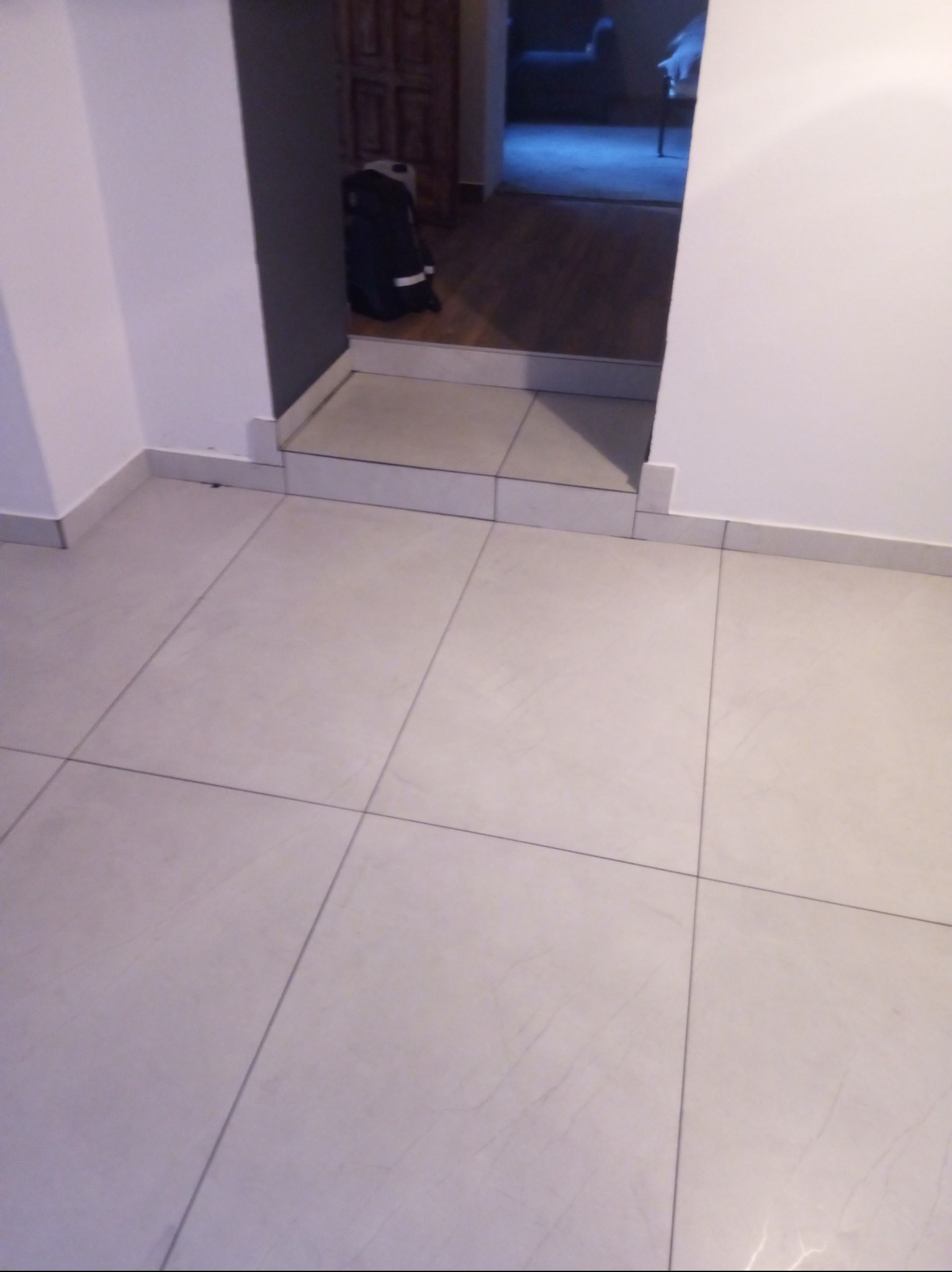 Wykończona podłoga z jasnych, kwadratowych płytek w przejściu między pomieszczeniami. Widoczna fuga i listwa przypodłogowa. W tle widoczny plecak.