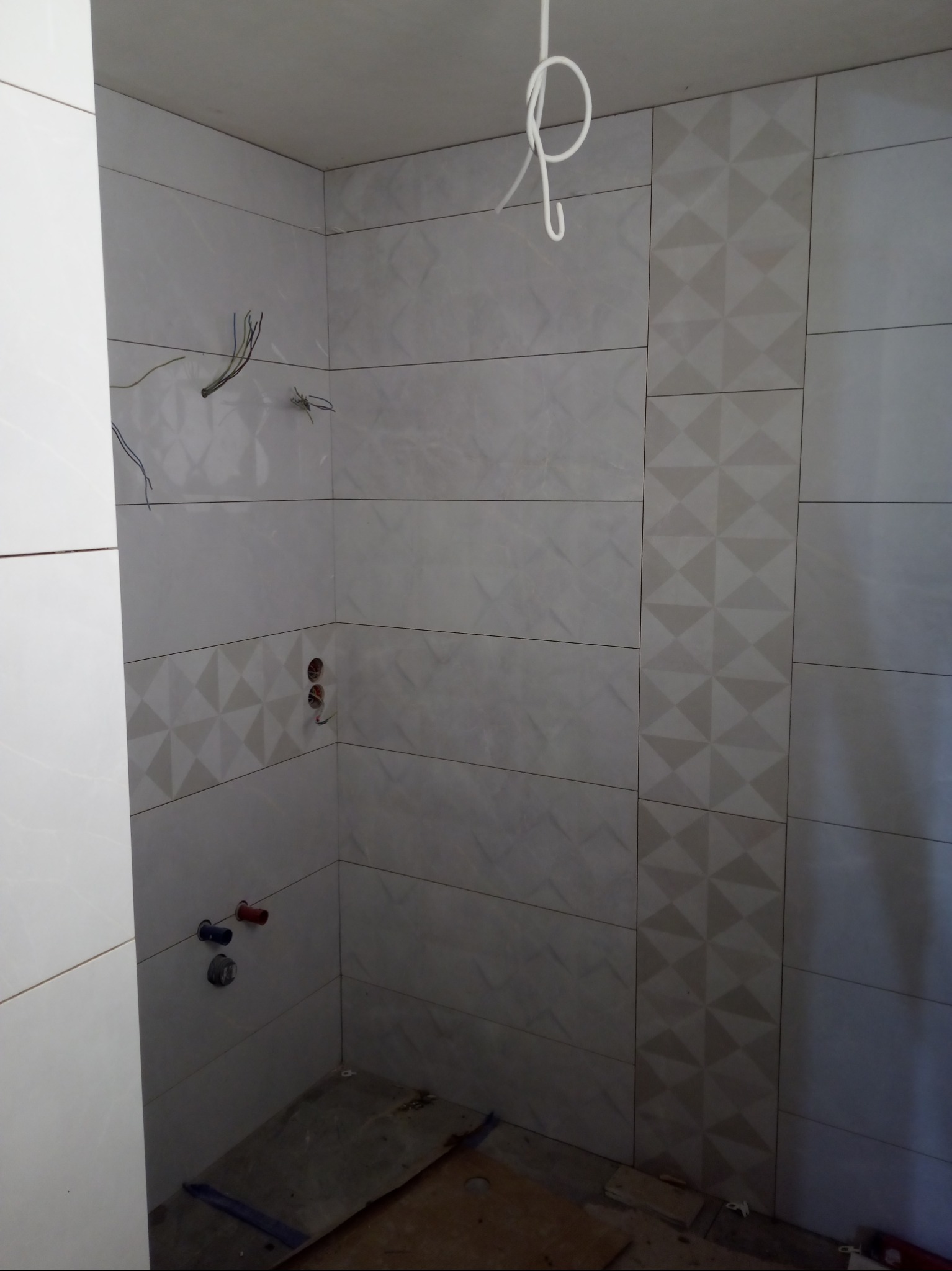 Ściany w łazience wyłożone jasnymi płytkami z geometrycznym wzorem. Widoczne wystające przewody i rury. Podłoga w trakcie prac wykończeniowych.