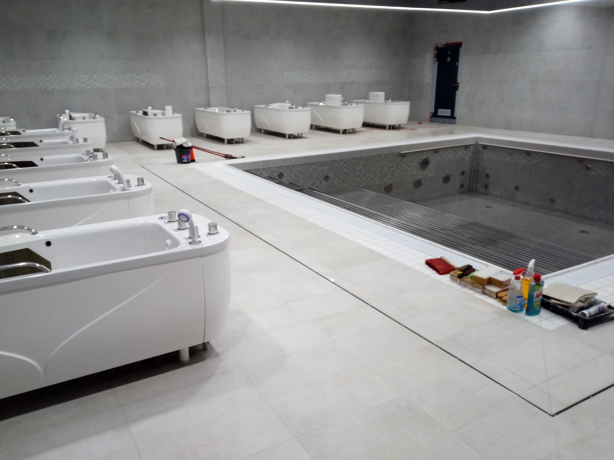 Wnętrze spa z rzędem białych wanien z hydromasażem i basenem wyłożonym mozaiką. Widoczne narzędzia i środki czystości, co sugeruje prace wykończeniowe lub sprzątanie.