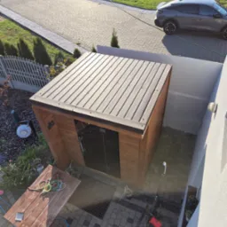 Nowoczesna, drewniana altana z metalowym dachem w ogrodzie, widziana z góry. Obok zaparkowany samochód i narzędzia ogrodowe na brukowanej powierzchni.