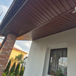 Radek Rybak - Brunatna podbitka dachowa z rynnami, widok z dołu na fragment elewacji z oknem i ceglanym filarem. W tle zieleń i zabudowa jednorodzinna.