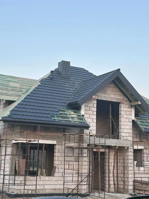 Dom w trakcie budowy z częściowo pokrytym dachem ciemną dachówką. Widoczne rusztowania i surowy stan ścian. Fragment dachu z zieloną membraną.