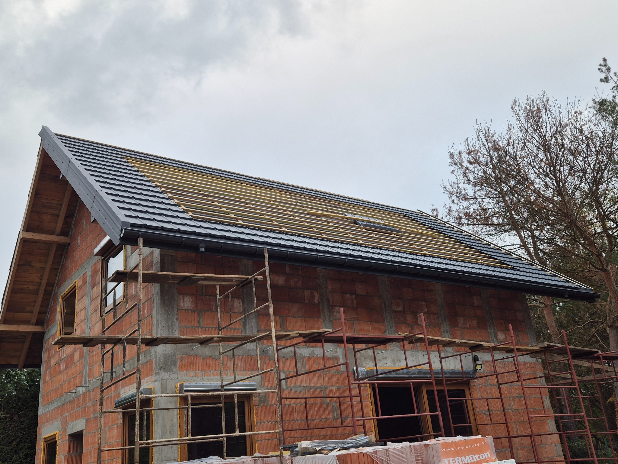 Budowa domu jednorodzinnego w trakcie. Widoczny fragment dachu z czarną dachówką i drewnianą konstrukcją, ściany z czerwonej cegły oraz rusztowanie.