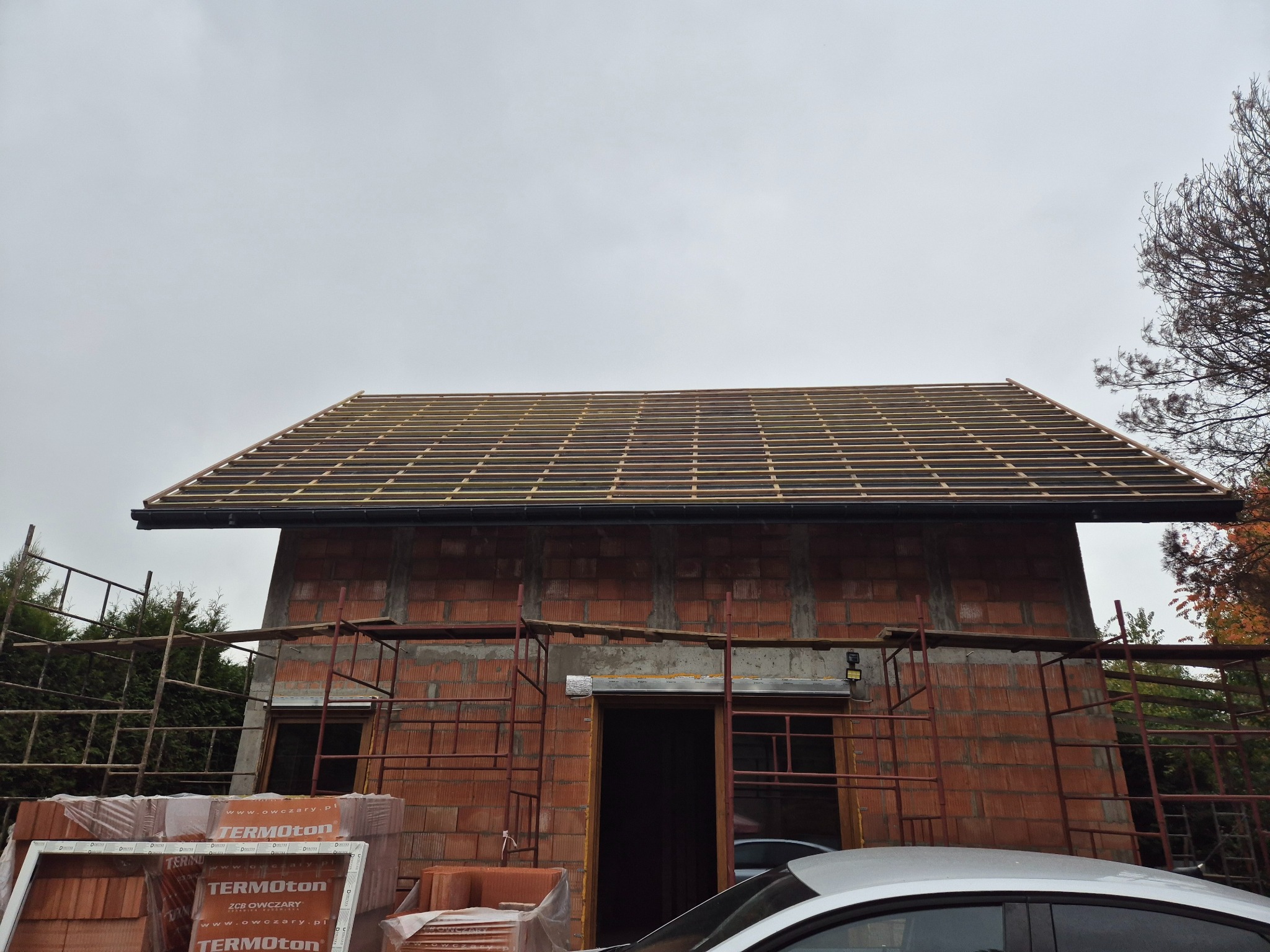 Budowa domu z widocznym szkieletem dachu z łat i kontrłat, częściowo pokrytym papą. Na pierwszym planie palety z bloczkami Termoton i fragment samochodu.