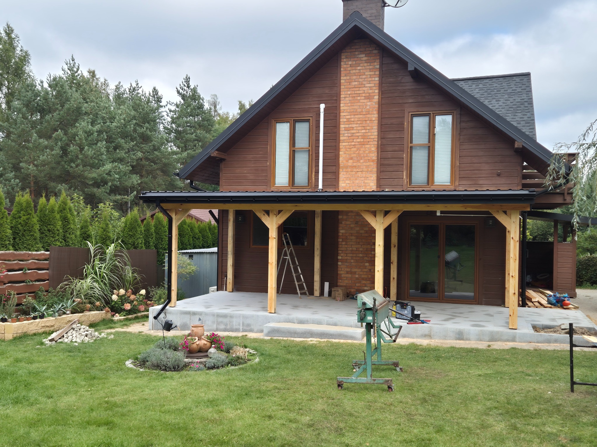 Dom z ciemnego drewna z nowym dachem z szarych dachówek bitumicznych. Widoczne narzędzia i elementy konstrukcyjne na tarasie. Tło: zielony trawnik i drzewa.