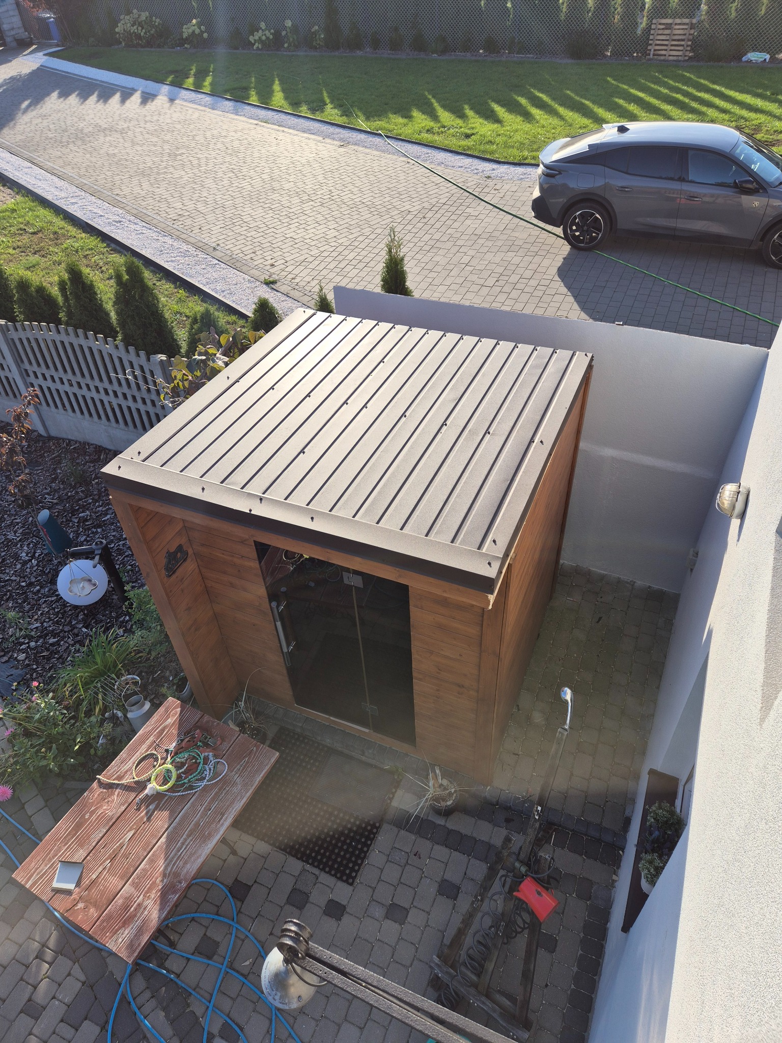 Nowoczesna, drewniana altana z metalowym dachem w ogrodzie, widziana z góry. Obok zaparkowany samochód i narzędzia ogrodowe na brukowanej powierzchni.