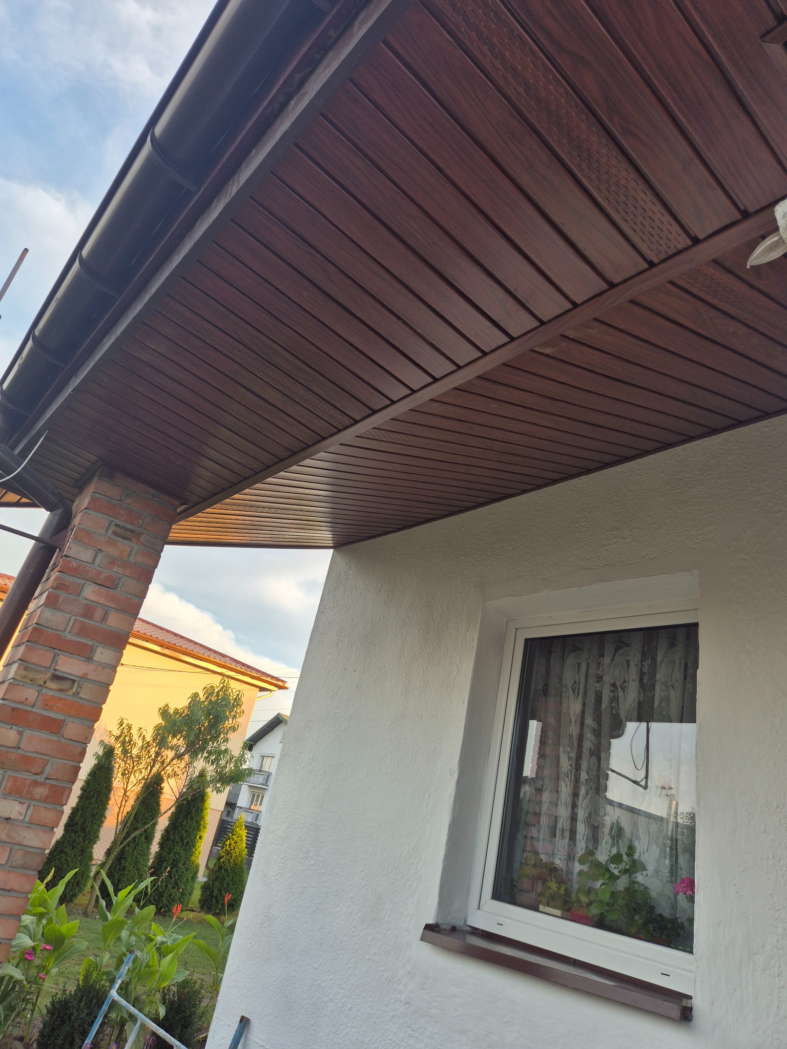 Brunatna podbitka dachowa z rynnami, widok z dołu na fragment elewacji z oknem i ceglanym filarem. W tle zieleń i zabudowa jednorodzinna.