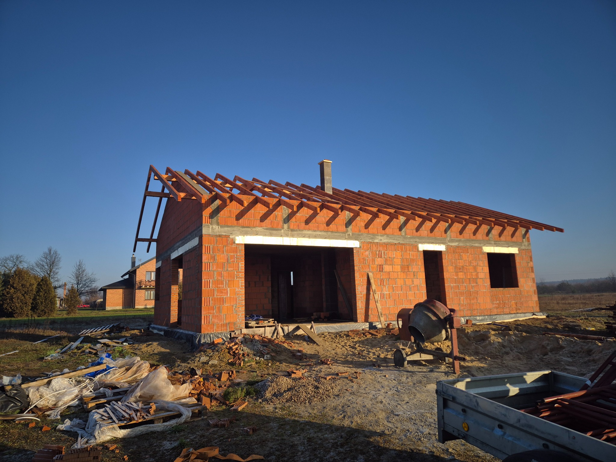 Surowy stan domu z czerwonej cegły z drewnianą konstrukcją dachu w trakcie budowy, widoczna betoniarka i przyczepa. W tle drugi budynek i błękitne niebo.