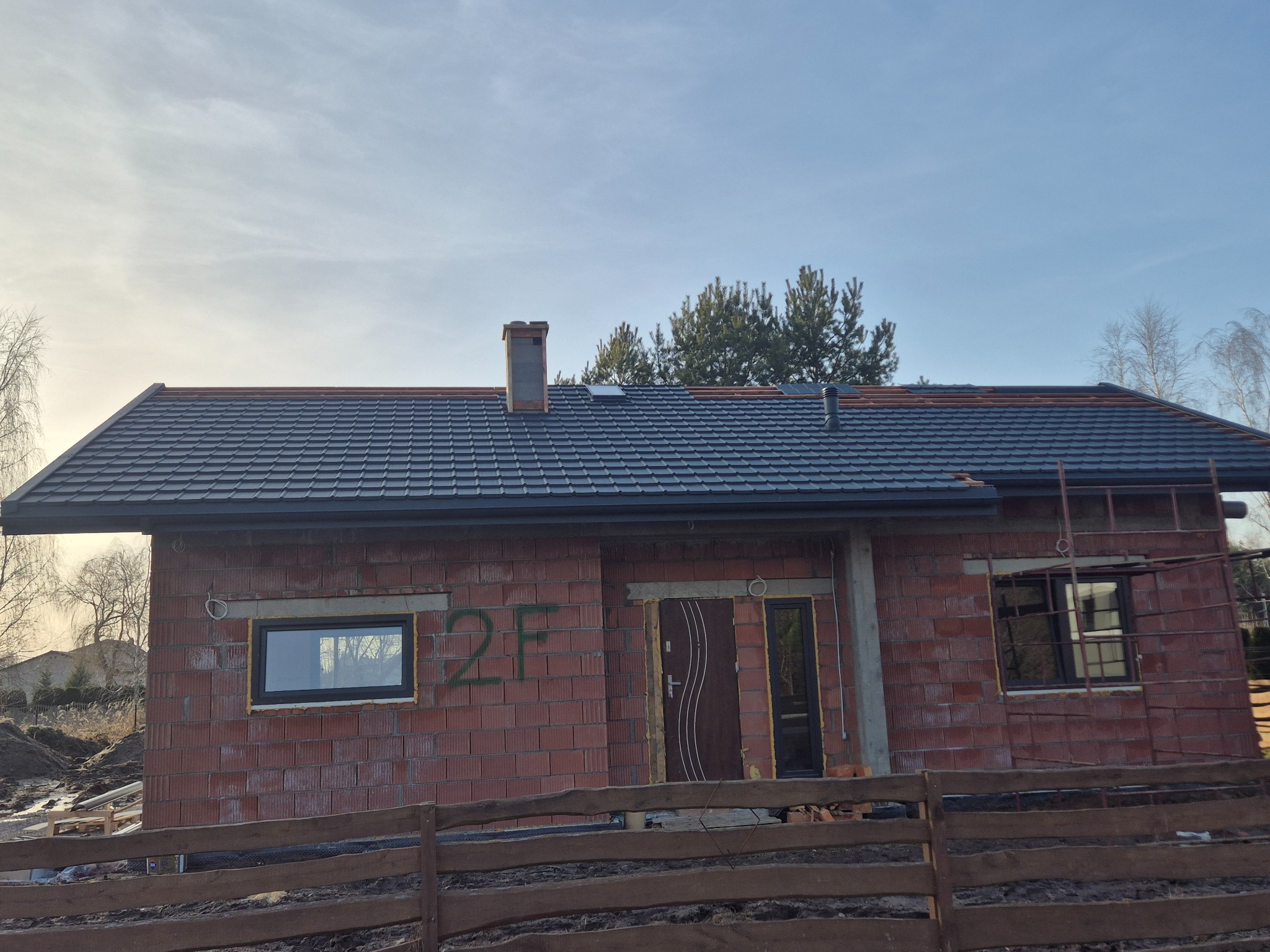 Dom w trakcie budowy z nowym, grafitowym dachem z blachodachówki. Widoczne rusztowanie, cegły i drewniane ogrodzenie. W tle drzewa i błękitne niebo.