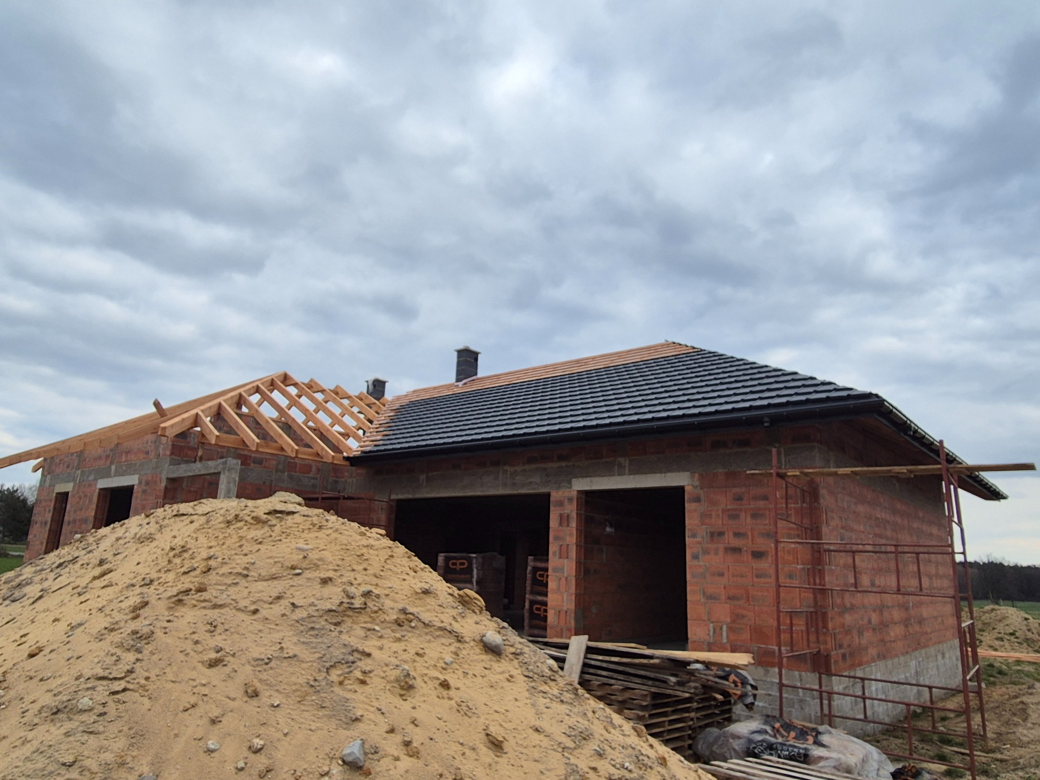 Budowa domu jednorodzinnego w trakcie: część dachu pokryta czarną dachówką, druga część z drewnianą konstrukcją. Na pierwszym planie hałda piasku.