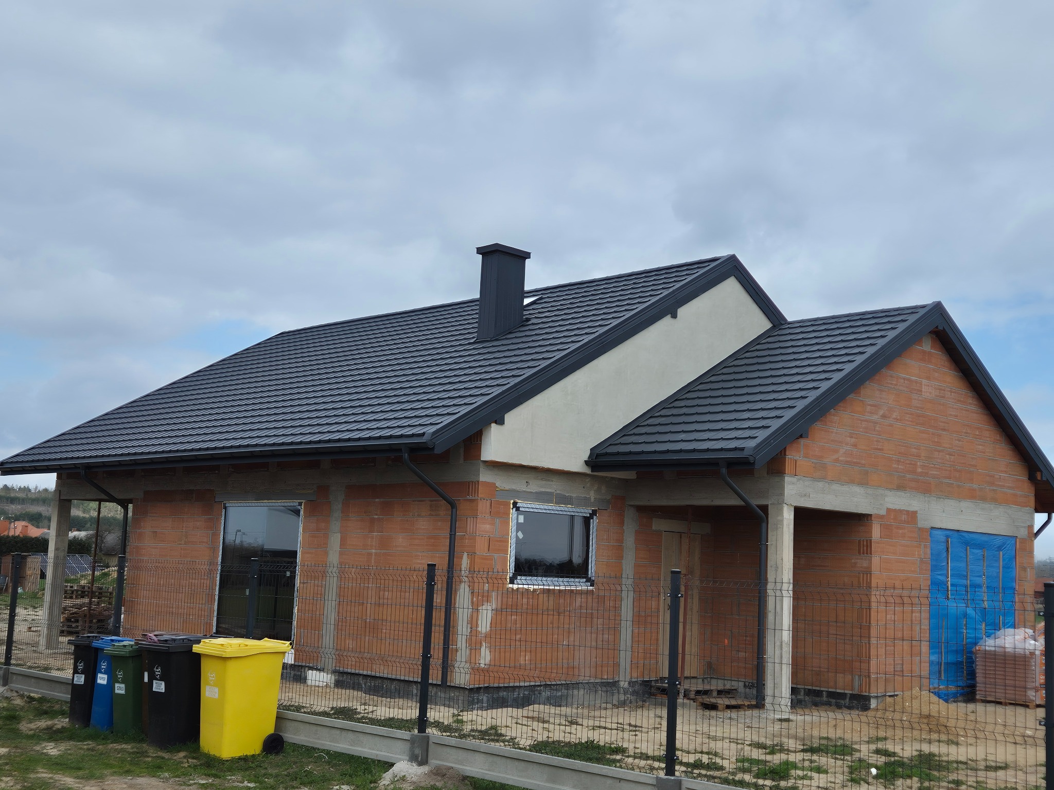 Dom w trakcie budowy z ciemnoszarą dachówką i kominem. Widoczne rynny, okna i ściany z cegły. Na pierwszym planie pojemniki na śmieci.