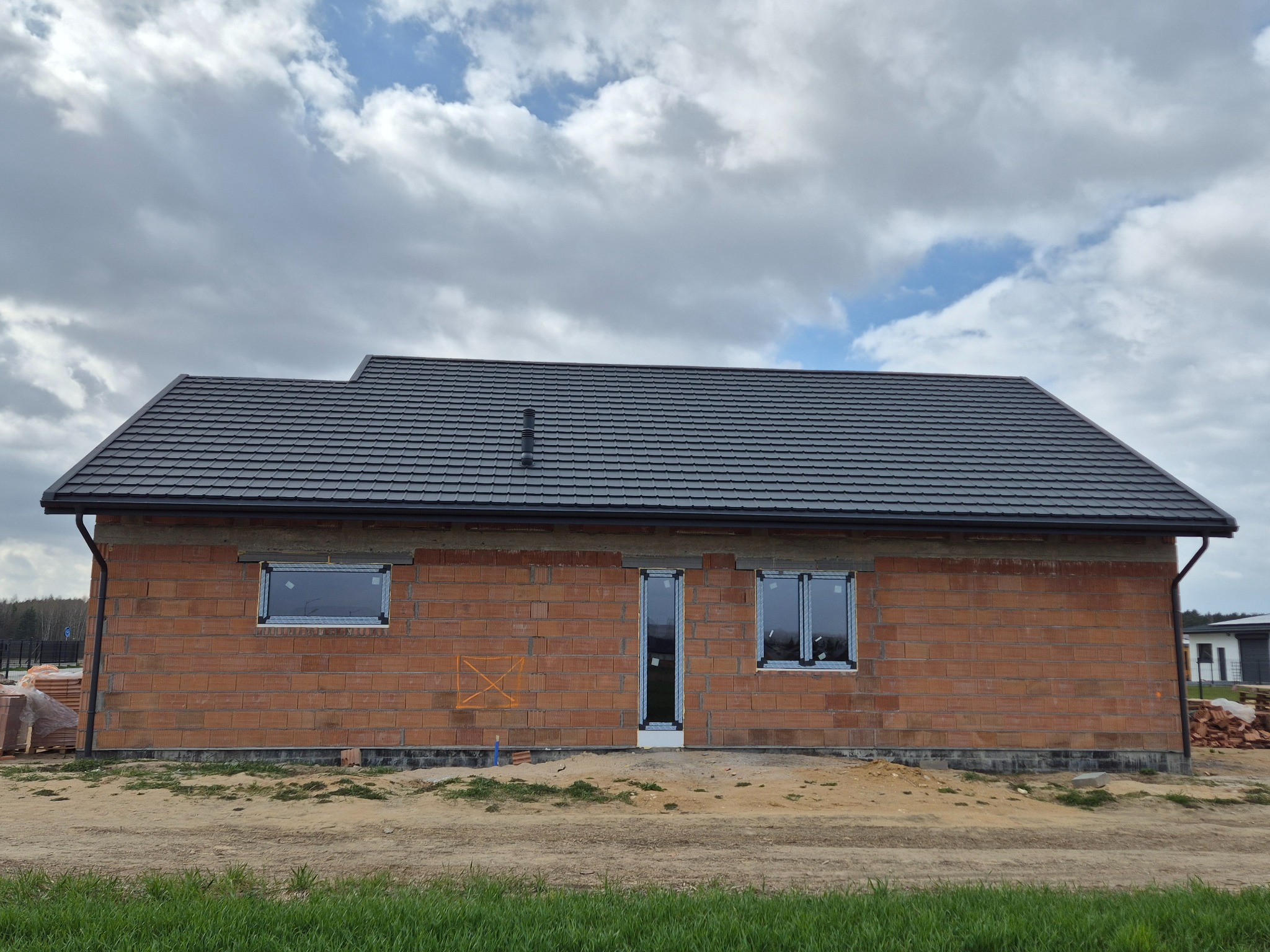 Dom w trakcie budowy z ciemnoszarym dachem z dachówki ceramicznej, widoczne okna i ceglana ściana. Na pierwszym planie trawa i ziemia.