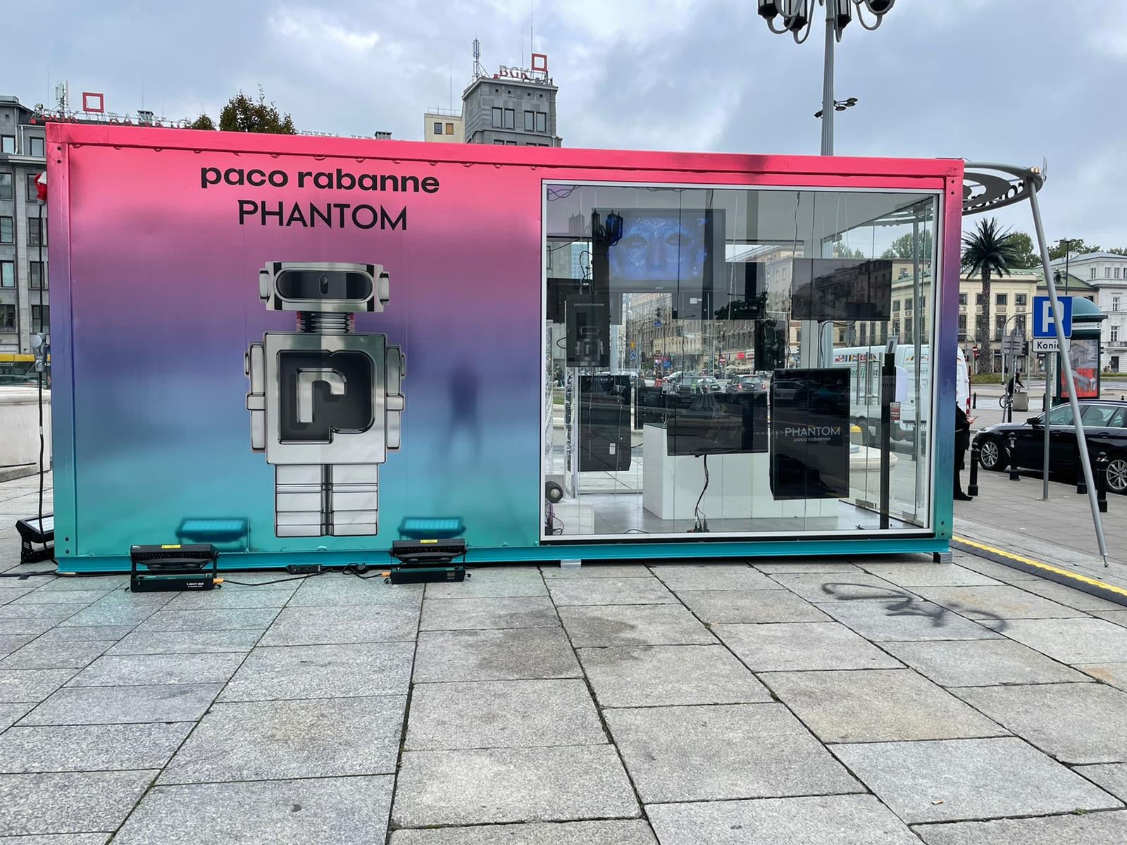 Kontener z grafiką Paco Rabanne Phantom, przeszklony, ustawiony na chodniku w mieście. Widoczne budynki w tle i oświetlenie kontenera. Intensywne kolory i nowoczesny design.