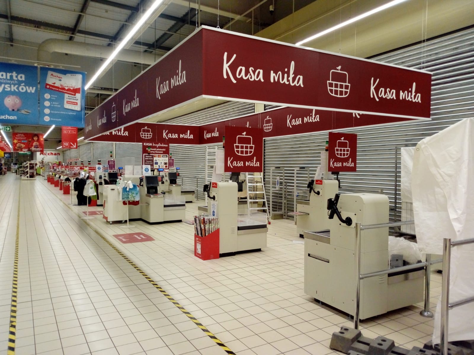 Oznakowanie strefy kas w supermarkecie: podwieszane, bordowe tablice z białym napisem 'Kasa miła' i grafiką koszyka. Widok na rzędy kas z zamkniętymi roletami.