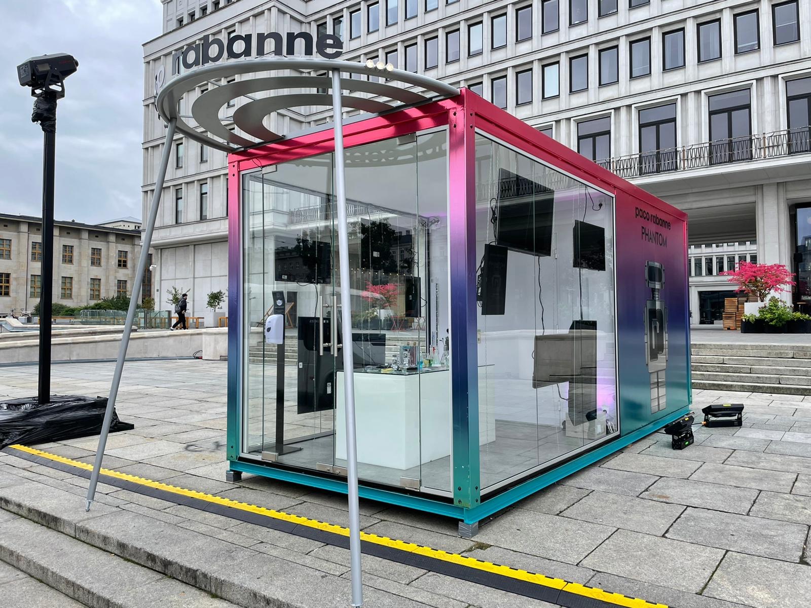 Paco Rabanne Phantom: kolorowy pawilon z logo na tle miejskiej architektury. Szklane ściany, wewnętrzne monitory, nowoczesny design, żółta linia bezpieczeństwa.