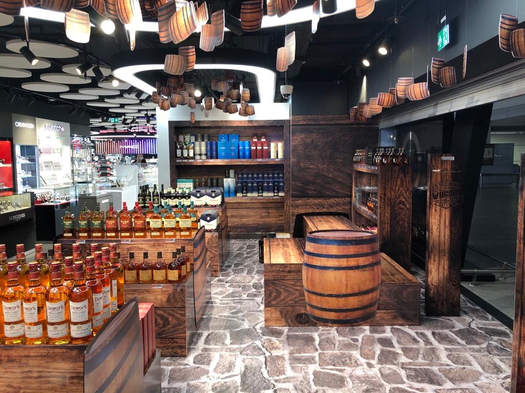 Aranżacja stoiska z alkoholem w stylu rustykalnym: beczki, drewno, kamienna podłoga, dekoracje z beczek pod sufitem. Produkty na półkach.