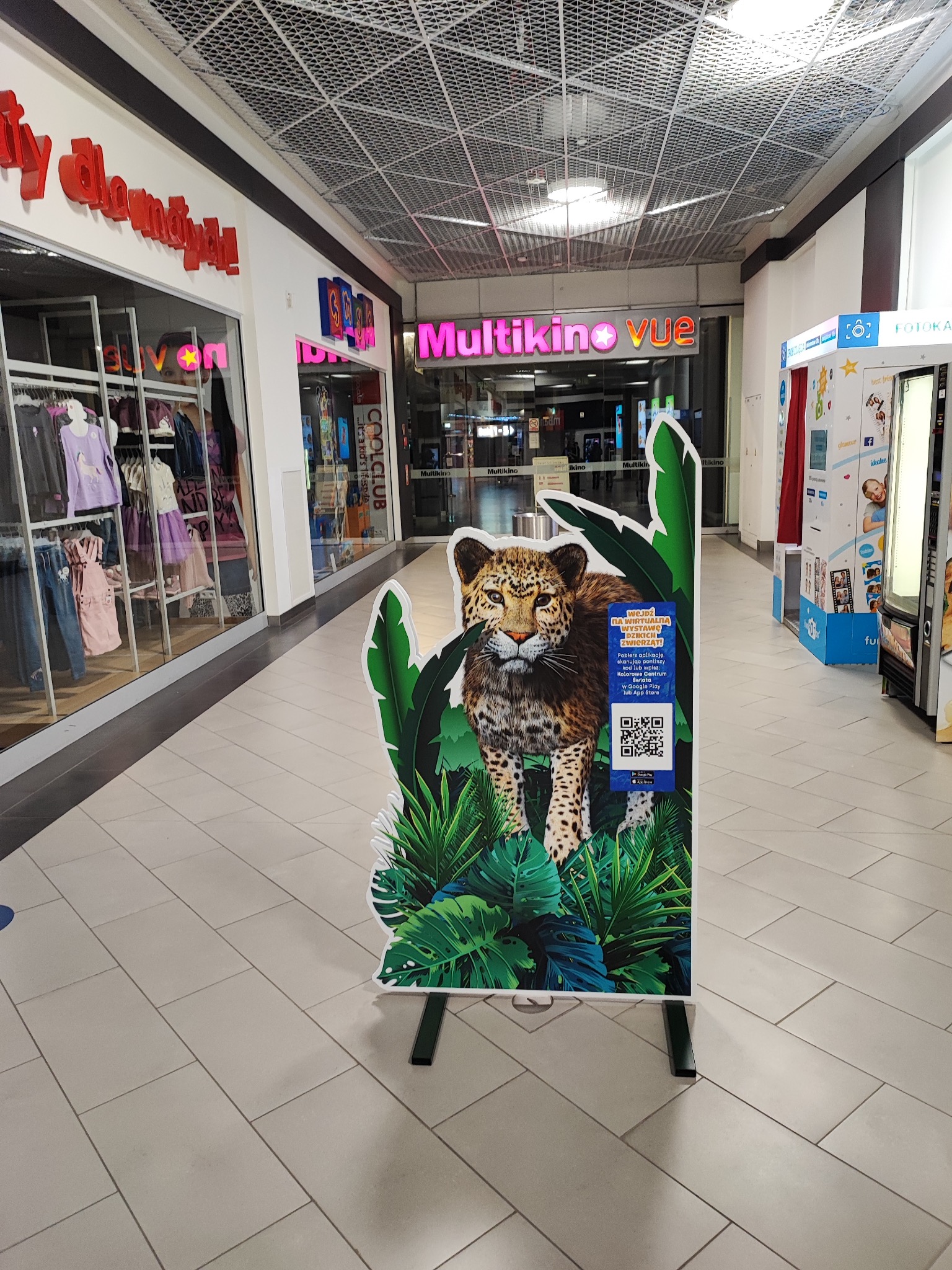 Stojak reklamowy z gepardem i tropikalną roślinnością w centrum handlowym, z kodem QR. W tle sklepy odzieżowe i Multikino.