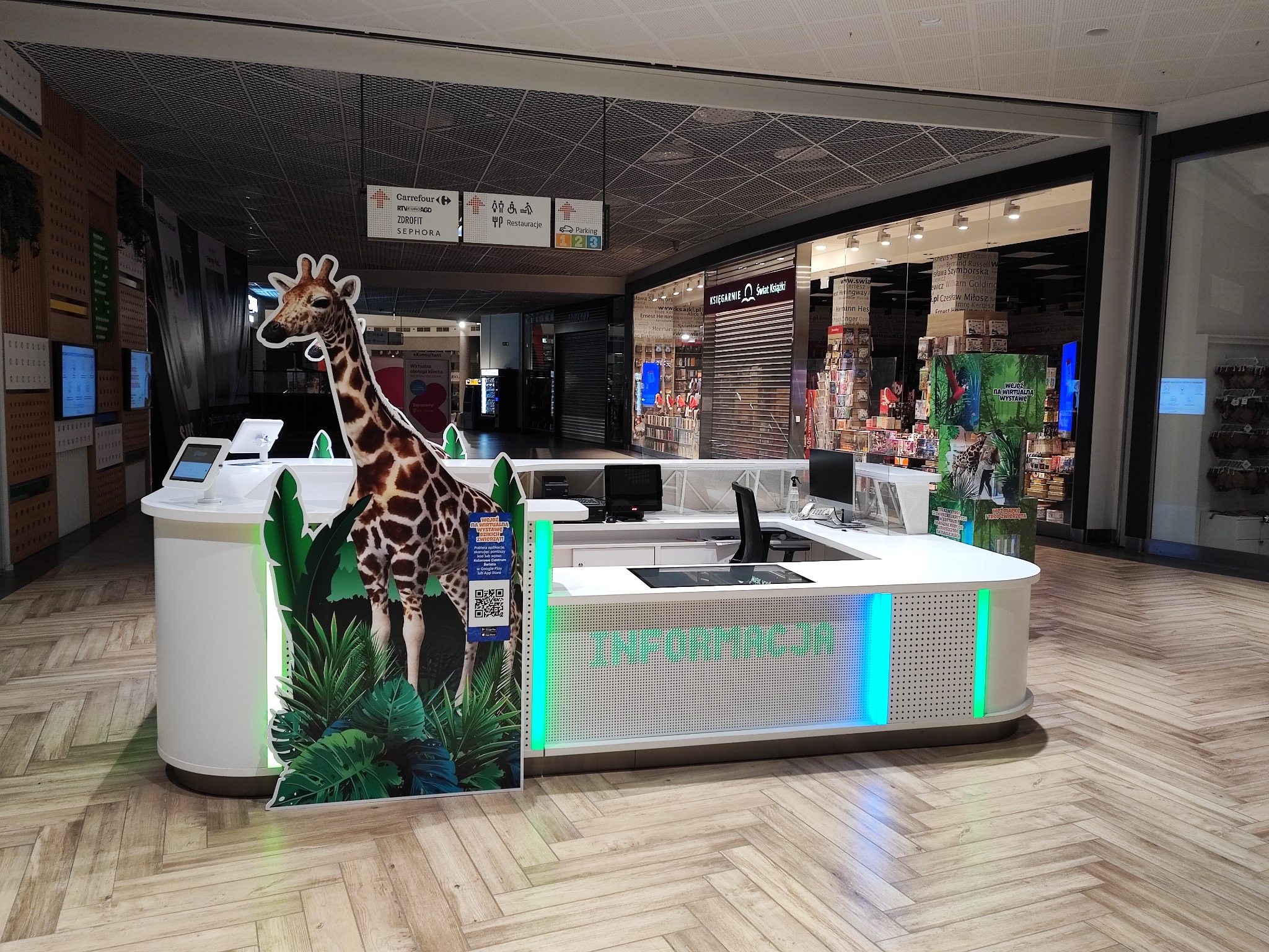 Stoisko informacyjne w galerii handlowej z grafiką żyrafy i podświetleniem LED, prezentujące wirtualną wystawę zwierząt.