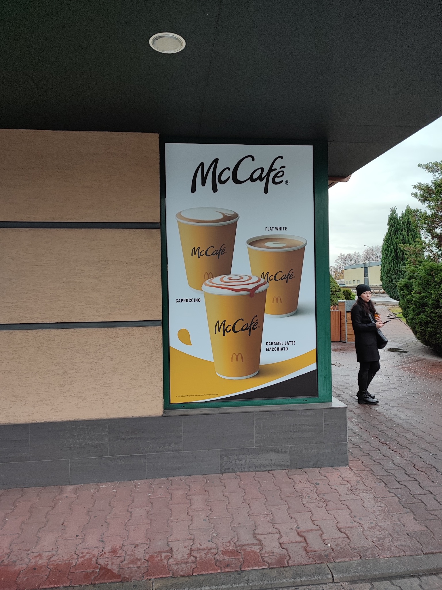 Reklama McCafé na zewnątrz budynku z kobietą w czarnym płaszczu. Plakat prezentuje cappuccino, flat white i caramel latte macchiato.