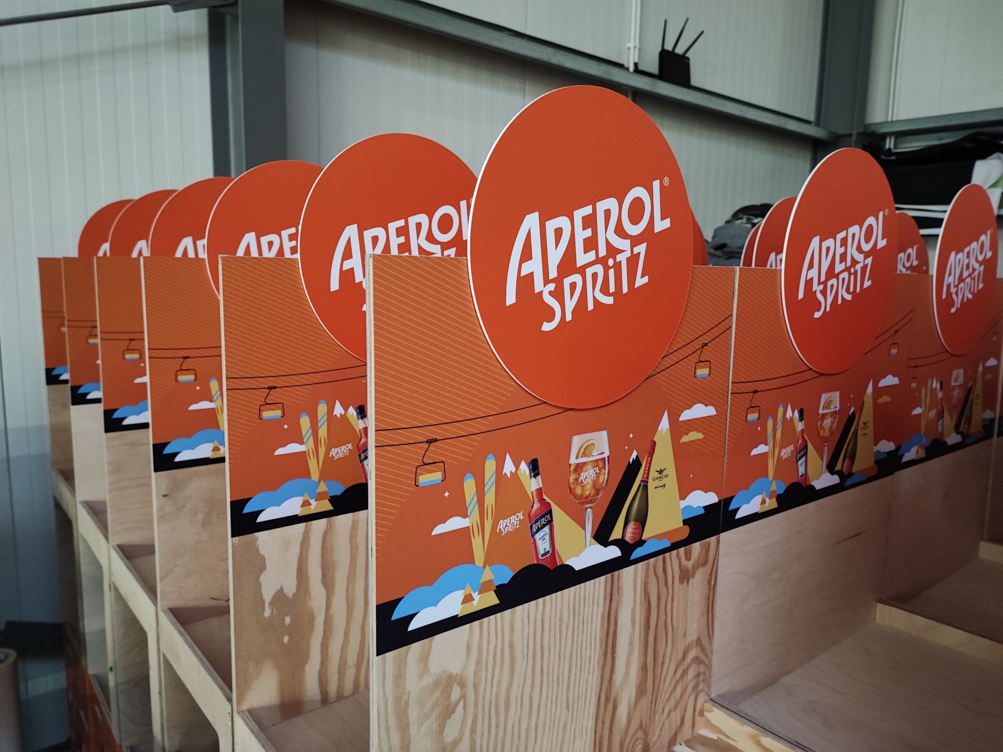 Rząd ekspozytorów Aperol Spritz z motywem zimowym, widoczne logo i grafika, wykonane w Warszawie. Konstrukcja z surowego drewna, pomarańczowe elementy.