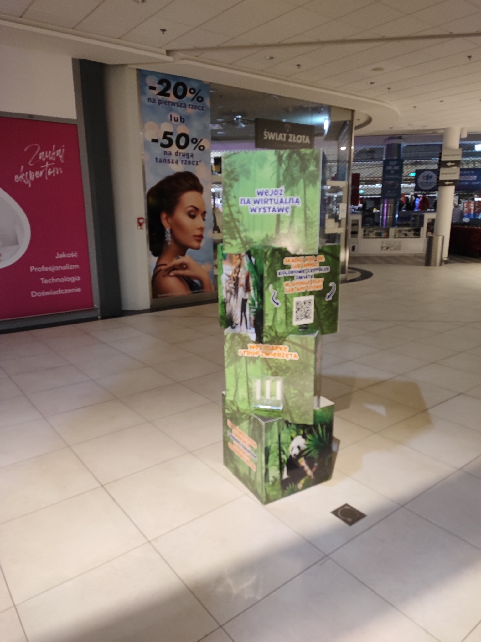 Wielopoziomowy, kartonowy stand reklamowy z grafiką zieleni i zwierząt, zachęcający do wejścia na wirtualną wystawę. W tle inne reklamy i elementy architektoniczne centrum handlowego.