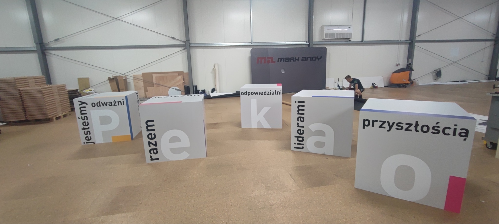 Kreatywne, białe, sześcienne konstrukcje z nadrukowanymi literami i hasłami w hali produkcyjnej. W tle widoczny pracownik i baner reklamowy.