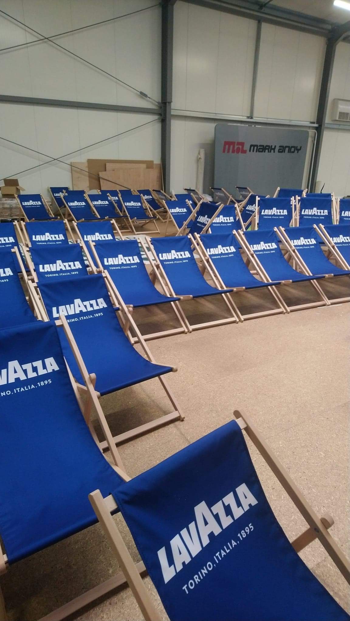 Rząd niebieskich leżaków z logo Lavazza w hali produkcyjnej. Widoczne logo Mark Andy w tle. Promocja marki na leżakach.