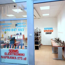 Wnętrze punktu naprawy elektroniki z ofertą: komputery, laptopy, tablety, konsole. Widoczne stanowiska serwisowe i półki z podzespołami. Oferta cenowa na szybie.