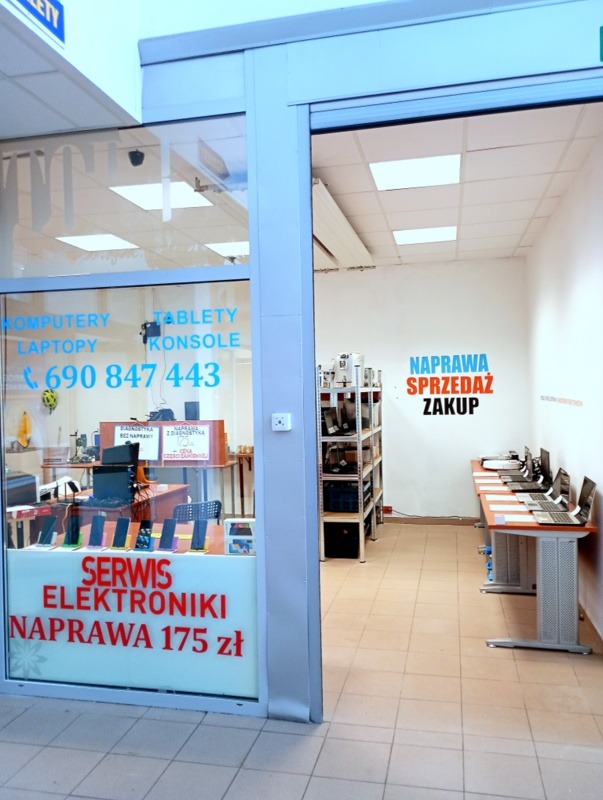 Wnętrze punktu naprawy elektroniki z ofertą: komputery, laptopy, tablety, konsole. Widoczne stanowiska serwisowe i półki z podzespołami. Oferta cenowa na szybie.