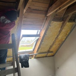 Expo-Activ - Ocieplenie poddasza wełną mineralną, widoczne drewniane elementy konstrukcyjne dachu i okno dachowe. Prace wykonywane z drabiny, narzędzia na podłodze.