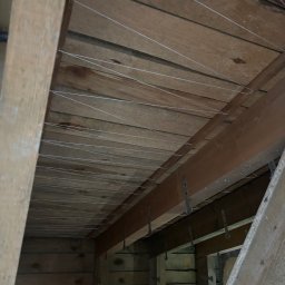 Expo-Activ - Drewniany strop z widocznymi elementami mocującymi i cienkimi przewodami, prawdopodobnie instalacja sufitu napinanego w trakcie montażu. Ujęcie z dołu.