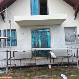 Expo-Activ - Elewacja domu w trakcie remontu: ocieplenie, rusztowania, okna zabezpieczone folią. Widoczne drewniane elementy dachu i fragment trawnika. Prace wykończeniowe.