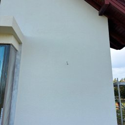 Expo-Activ - Ściana domu po nałożeniu jasnego tynku strukturalnego, widoczny fragment okna z folią ochronną, fragment dachu z brązową dachówką i rusztowanie z boku.