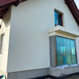 Expo-Activ - Elewacja domu jednorodzinnego po nałożeniu tynku, z zabezpieczonymi oknami folią ochronną. Widoczne detale wykończenia i elementy instalacji zewnętrznej.