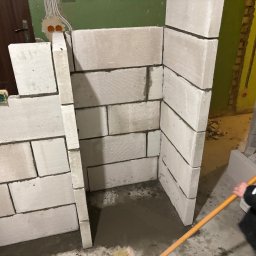 Expo-Activ - Wznoszenie ścianki działowej z bloczków betonu komórkowego w surowym wnętrzu. Widoczne narzędzia i zaprawa. Prace murarskie w trakcie realizacji.