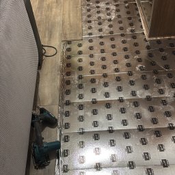 Expo-Activ - Izolacja podłogowa z folii aluminiowej z nadrukiem, częściowo ułożona na drewnianej podłodze, obok narzędzia do cięcia. Widok z góry, ukośny kadr.