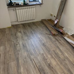 Expo-Activ - Układanie paneli podłogowych w pokoju. Częściowo ułożona podłoga z paneli w odcieniach brązu, paczki paneli przy ścianie, narzędzia na parapecie okna.