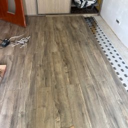 Expo-Activ - Ułożone panele podłogowe o wzorze drewna, widoczne narzędzia i podkład. Częściowo widoczna ościeżnica drzwi i szafa przesuwna. Podłoże przygotowane pod dalszy montaż.