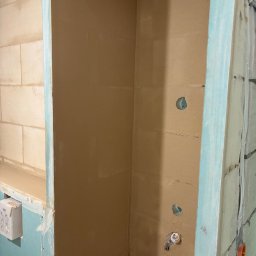 Expo-Activ - Ściana w trakcie szpachlowania z widocznymi otworami na instalacje, obudowana płytą gipsową z niebieskimi krawędziami. Fragment ściany z cegieł w tle.