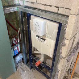Expo-Activ - Stelaż podtynkowy Grohe zamontowany w łazience, widoczne instalacje wodne i kanalizacyjne, ściany z bloczków, metalowa konstrukcja, etap prac wykończeniowych.
