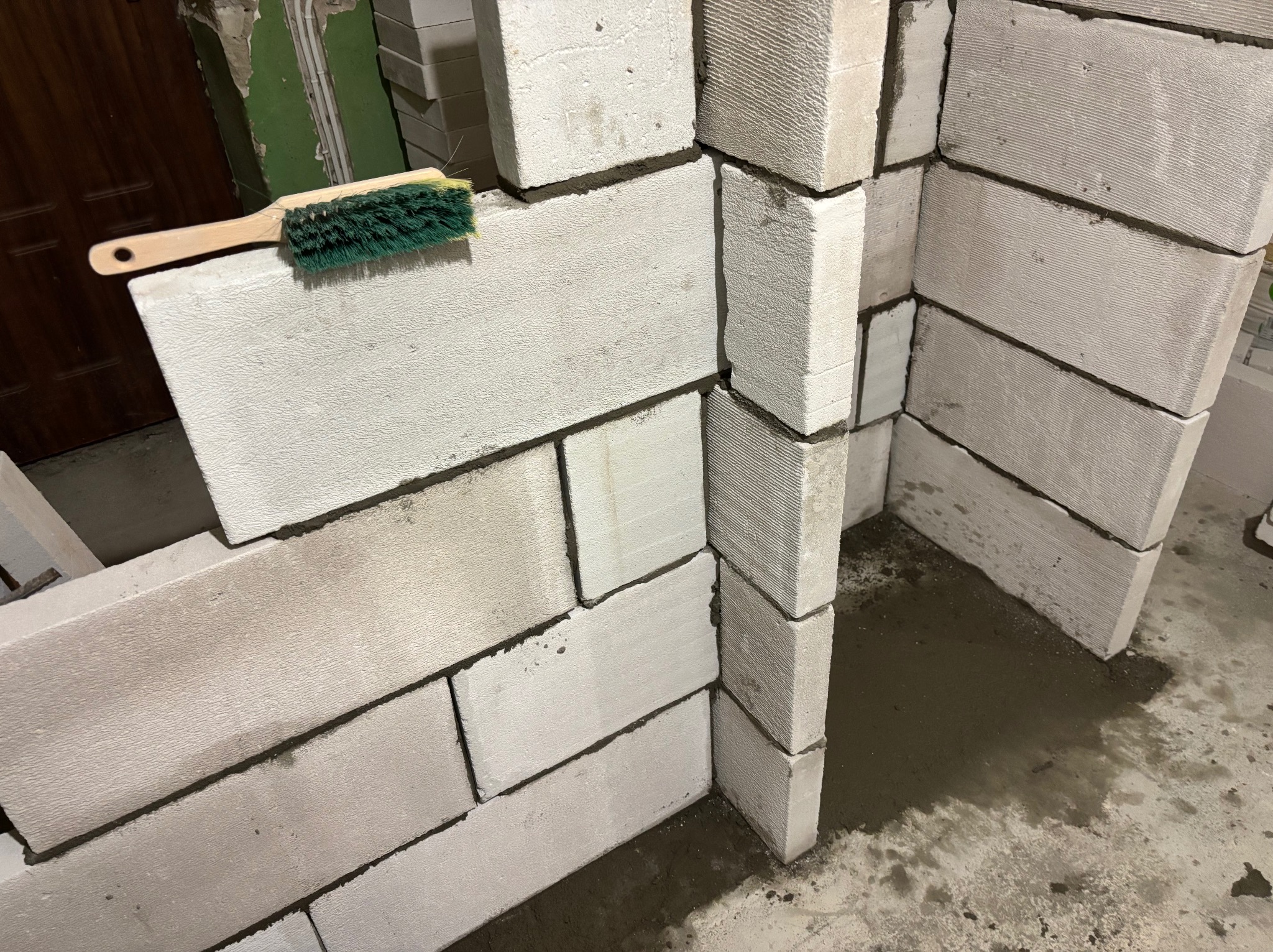 Wznoszenie ściany z białych bloczków betonu komórkowego, widoczna szczotka do czyszczenia spoin, surowy stan wykończenia podłogi i ściany w tle.