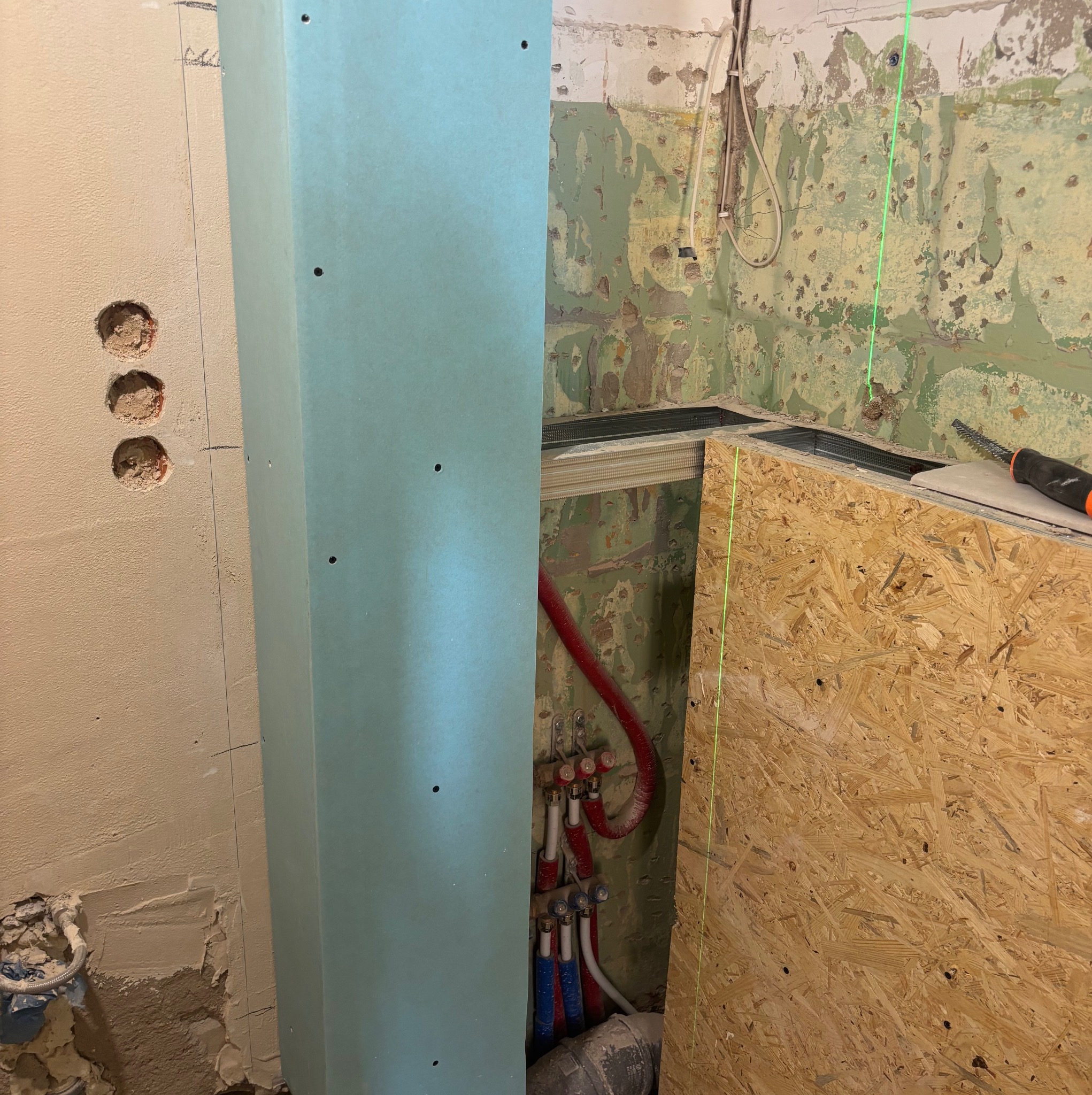 Ściana z płyt karton-gips, widoczne instalacje wodne i elektryczne, surowy stan przed wykończeniem. Laserowy niwelator wyznacza pion. Fragment płyty OSB w tle.