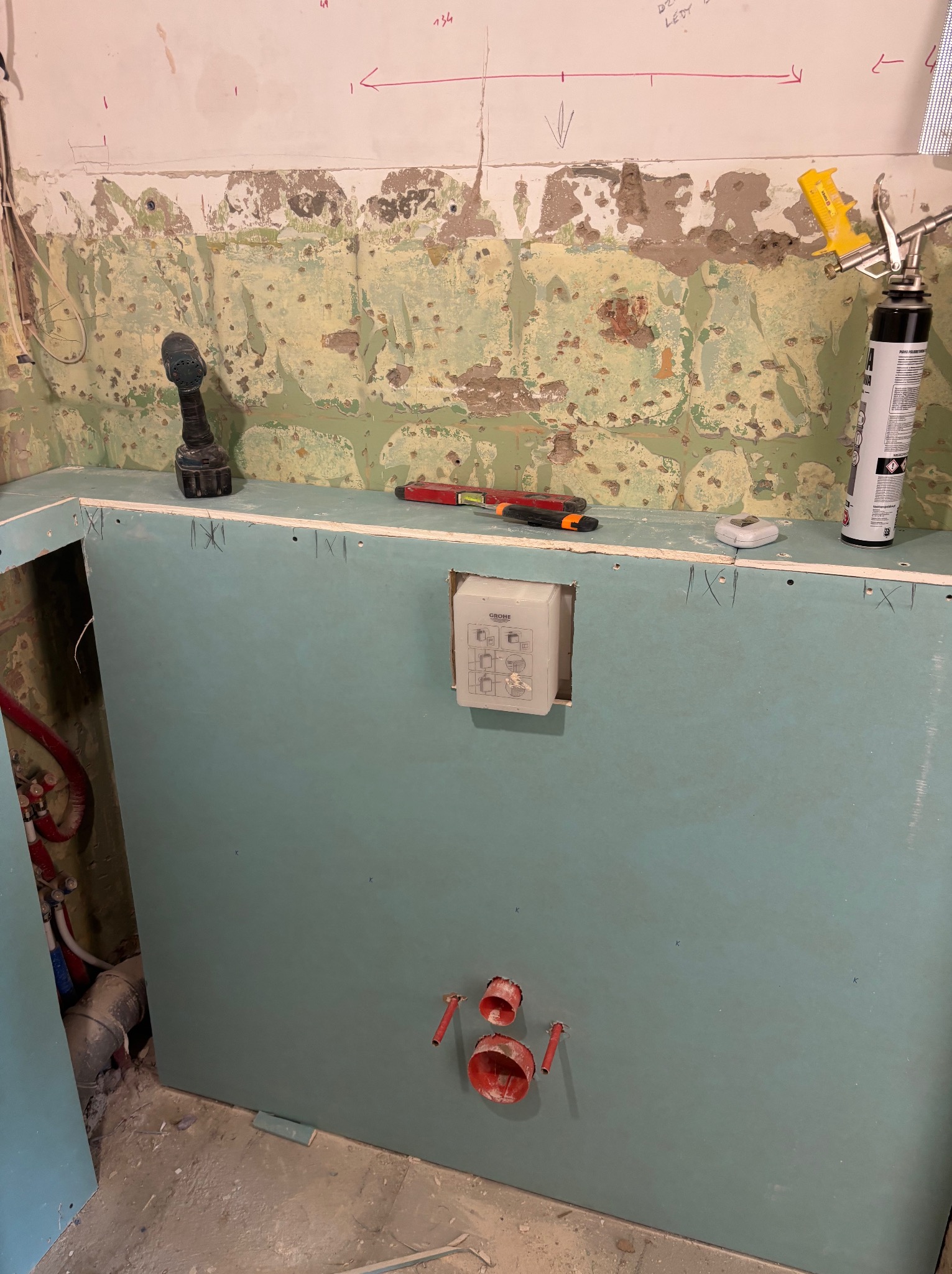 Ściana z płyty gipsowo-kartonowej w trakcie montażu w łazience. Widoczne narzędzia, instalacja wodna i fragment starej ściany z resztkami farby. Otwory na rury.