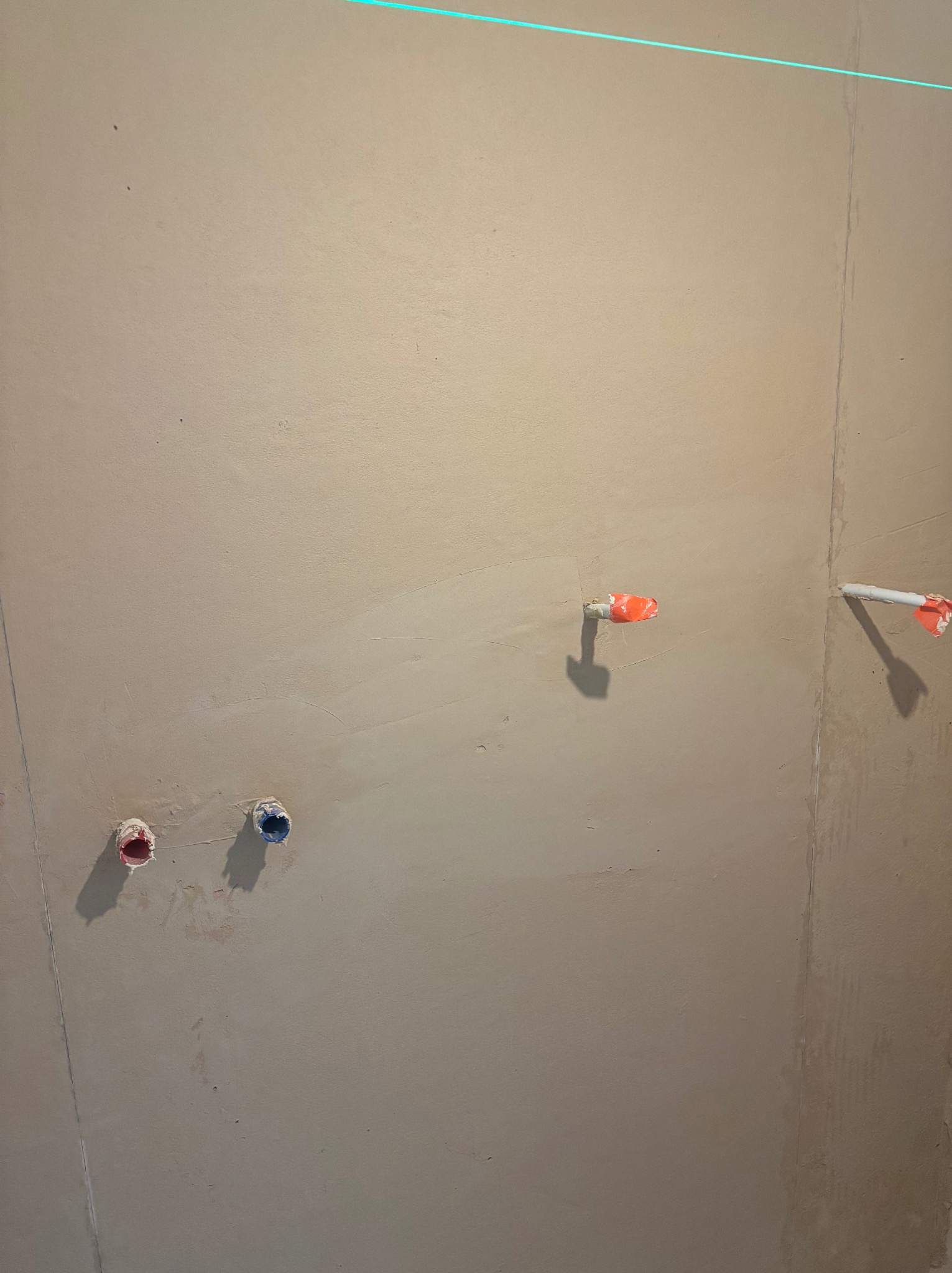 Ściana po tynkowaniu z wystającymi rurami instalacji wodnej zabezpieczonymi taśmą, widoczna linia lasera wyznaczająca poziom. Szare tło, surowy stan wykończenia.