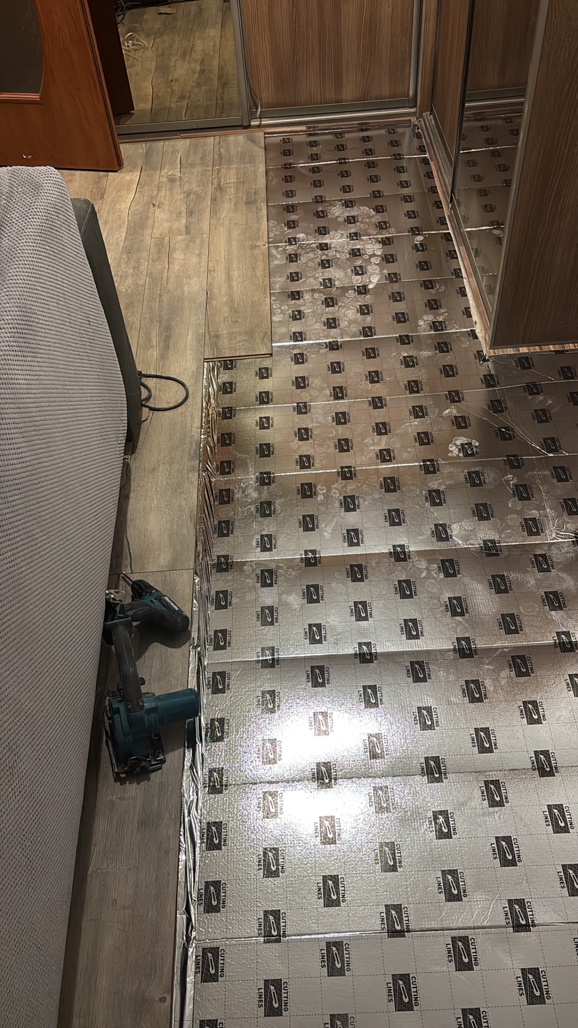 Izolacja podłogowa z folii aluminiowej z nadrukiem, częściowo ułożona na drewnianej podłodze, obok narzędzia do cięcia. Widok z góry, ukośny kadr.